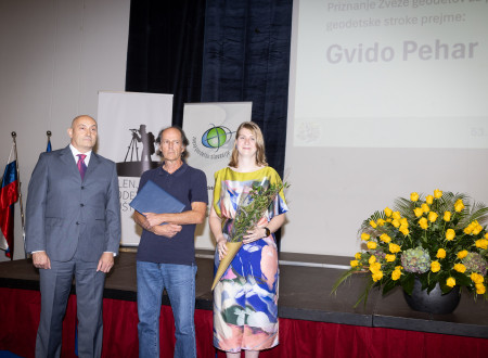 Moški stoji z listino med podeljevalcema; v ozadju sta zastavi in logotipa ZGS ter Dolenjskega geodetskega društva, ob strani je cvetlični aranžma.