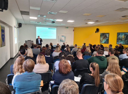 Moški predavatelj stoji pred občinstvom v dvorani z rumeno steno in projiciranim programom strokovnega posveta.