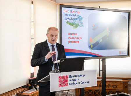 Moški v poslovni obleki govori za govorniškim pultom; na zaslonu poleg njega je diapozitiv o horizontalnem povezovanju in krožni ekonomiji prostora.