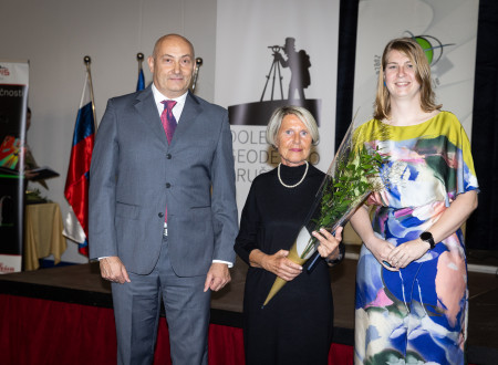 Ženska s šopkom stoji med podeljevalcema na odru; v ozadju sta zastavi in logotipa ZGS ter Dolenjskega geodetskega društva.