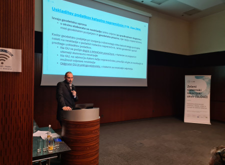 Moški predavatelj stoji za govorniškim pultom pred projekcijskim platnom z besedilom o uskladitvi podatkov katastra nepremičnin, ob strani je pano SLO4D.