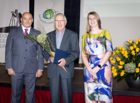 Moški s šopkom in listino stoji med predstavnikoma podeljevalca; v ozadju sta zastavi in logotipa ZGS ter Dolenjskega geodetskega društva in projekcija, ob strani je cvetlični aranžma.