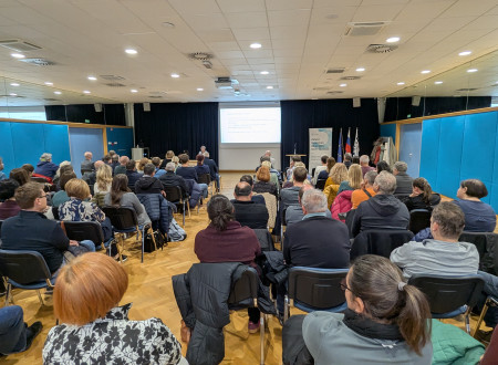 Ženska predavateljica stoji za govorniškim pultom pred projekcijskim platnom s programom posveta ter ob panoju SLO4D in državni zastavi.