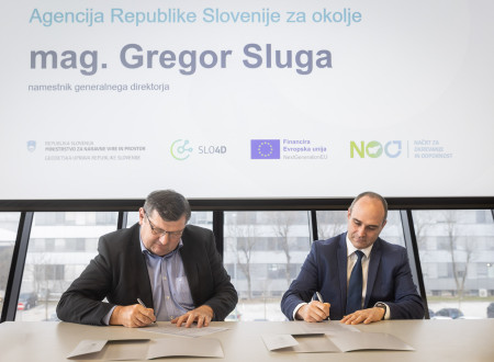 Dva moška sedita za mizo in kot predstavnika Agencije RS za okolje ter Geodetske uprave RS podpisujeta dokumente o skupnosti Geo Slovenija, v ozadju je projekcija z imenom mag. Gregorja Sluge.
