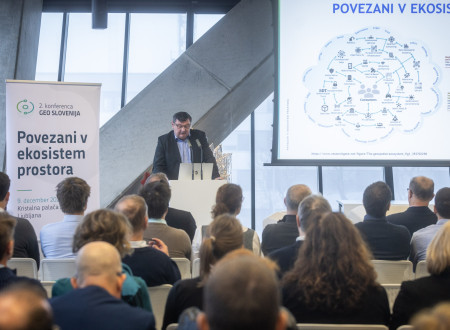 V dvorani Kristalne palače moški govorec stoji za govorniškim pultom in pred polno zasedeno publiko predstavlja diagram povezanega ekosistema prostora, ob njem pa stoji pano 2. konference Geo Slovenija.