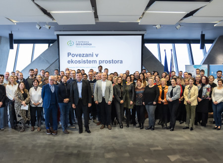 Velika skupina udeležencev različnih starosti stoji v več vrstah v konferenčni dvorani pred platnom z napisom »Povezani v ekosistem prostora« in se ozira proti fotoaparatu.