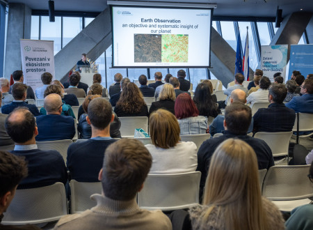 Moški govorec pred polno dvorano na velikem platnu prikazuje diapozitiv z napisom »Earth Observation« in dvema satelitskima posnetkoma površja, udeleženci sedijo v vrstah in spremljajo predstavitev.