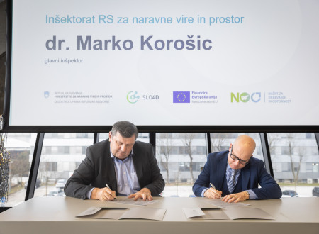 Dva moška sedita za mizo in kot predstavnika Inšpektorata RS za naravne vire in prostor ter Geodetske uprave RS podpisujeta dokumente pred projekcijskim platnom z imenom govorca.