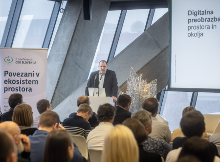 Moški govorec v obleki stoji za govorniškim pultom pred občinstvom, na projekcijskem platnu pa je prikazan diapozitiv z naslovom »Digitalna preobrazba prostora in okolja« in pano 2. konference Geo Slovenija.