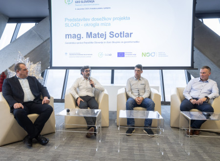 Štirje moški sedijo v bež foteljih pred projekcijskim platnom, eden govori v mikrofon, ostali poslušajo, za njimi pa je diapozitiv z naslovom okrogle mize o dosežkih projekta SLO4D.
