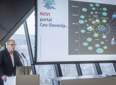 Moški govorec stoji za govorniškim pultom in ob projekcijskem platnu, na katerem je prikazan koncept novega portala Geo Slovenija z mrežo krožnih ikon, občinstvo sedi in spremlja predavanje.