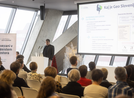 Moški govorec stoji pred projekcijskim platnom z diagramom »Kaj je Geo Slovenija?«, v dvorani pa sedi več vrst udeležencev, ki spremljajo predstavitev koordinacijskega centra in delovnih skupin.
