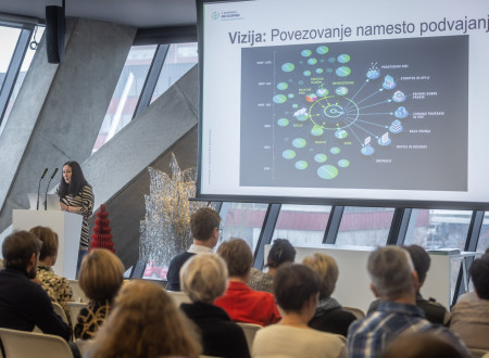 Ženska govorka stoji za govorniškim pultom in predavateljsko publiko, na platnu pa je prikazan grafični prikaz vizije »Povezovanje namesto podvajanja« z med seboj povezanimi tematskimi sklopi.