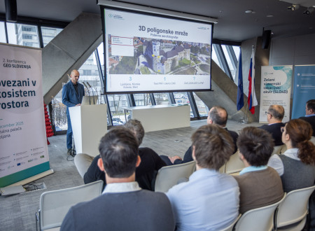 Moški govorec stoji pri beli mizi z računalnikom, na projekcijskem platnu pa je prikazan 3D prikaz gradu in napis o 3D poligonskih mrežah, občinstvo v prvih vrstah spremlja predstavitev.