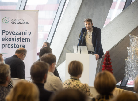 Moški govorec stoji za govorniškim pultom ob panoju »Povezani v ekosistem prostora«, pred njim pa sedi več vrst udeležencev, ki poslušajo predstavitev o geodetskih podatkih in tehnologijah.
