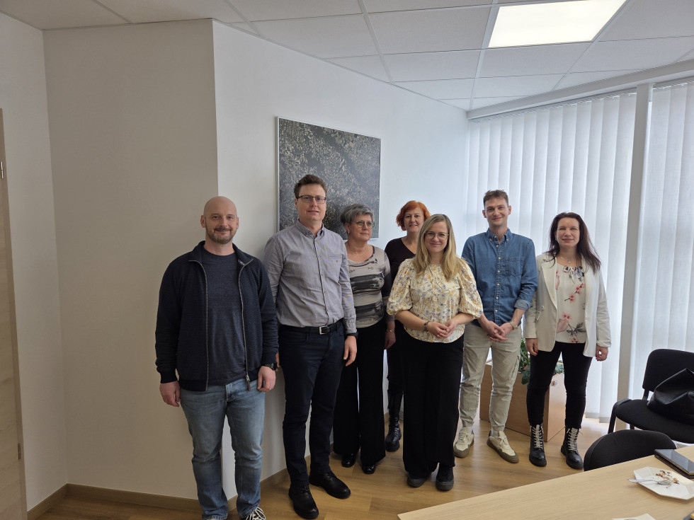 V prostoru – pisarni stojijo skupaj štiri ženske in trije moški. V ozadju je na steni vidna slika ki prikazuje posnetek mesta.