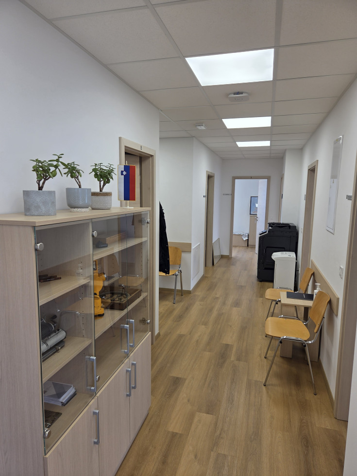 V prostoru - hodniku, so vidni trije stoli za obiskovalce, mizica ter omara, na kateri so trije lonci z rožami. Globje v hodniku stoji kopirni stroj.