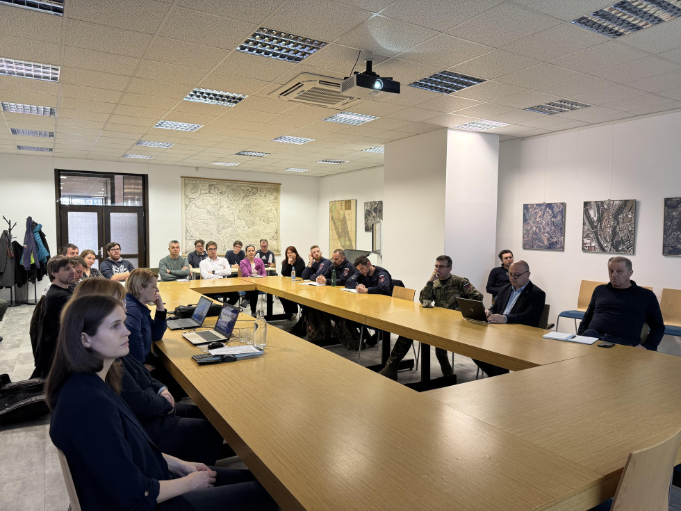 V sejni sobi za mizo sedijo osebe, moški in ženske, ki zbrano poslušajo predstavitev. Omizje je sklenjeno v pravokotni obliki, na stenah vidijo slike.