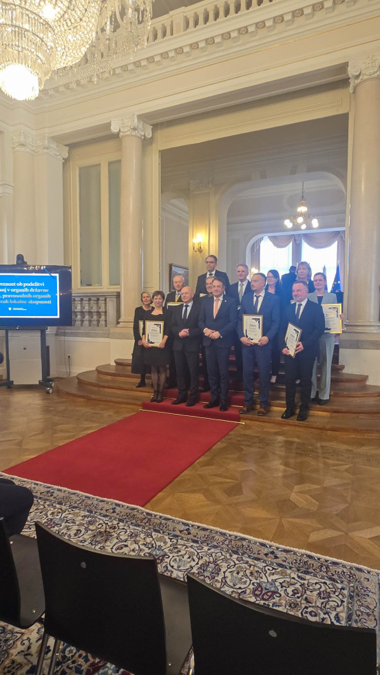 Skupina moških in žensk stoji na stopnicah v veliki dvorani Predsedniške palače. V rokah držijo plakete, v ozadju so stebri, lestenec in slavnostni prostor za podelitev priznanj.