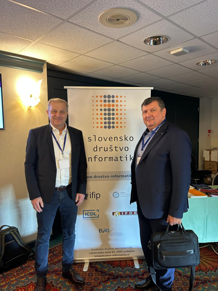 Dva moška v poslovni obleki stojita pred promocijskim panojem Slovenskega društva informatika, eden drži poslovno torbo, za njima je razstavni prostor konference.