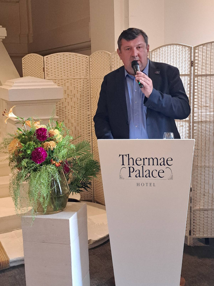 Moški v temni obleki stoji za govorniškim pultom hotela Thermae Palace in govori v mikrofon med uradnim dogodkom.