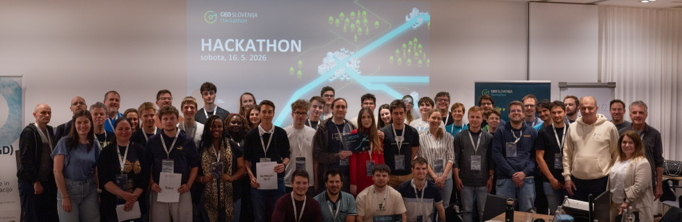 Večja skupina moških in žensk stoji v konferenčni dvorani pred projekcijo z napisom Hackathon. Nekateri udeleženci v rokah držijo priznanja za sodelovanje in nagrade, v ozadju je grafika Geo Slovenija Hackathona.