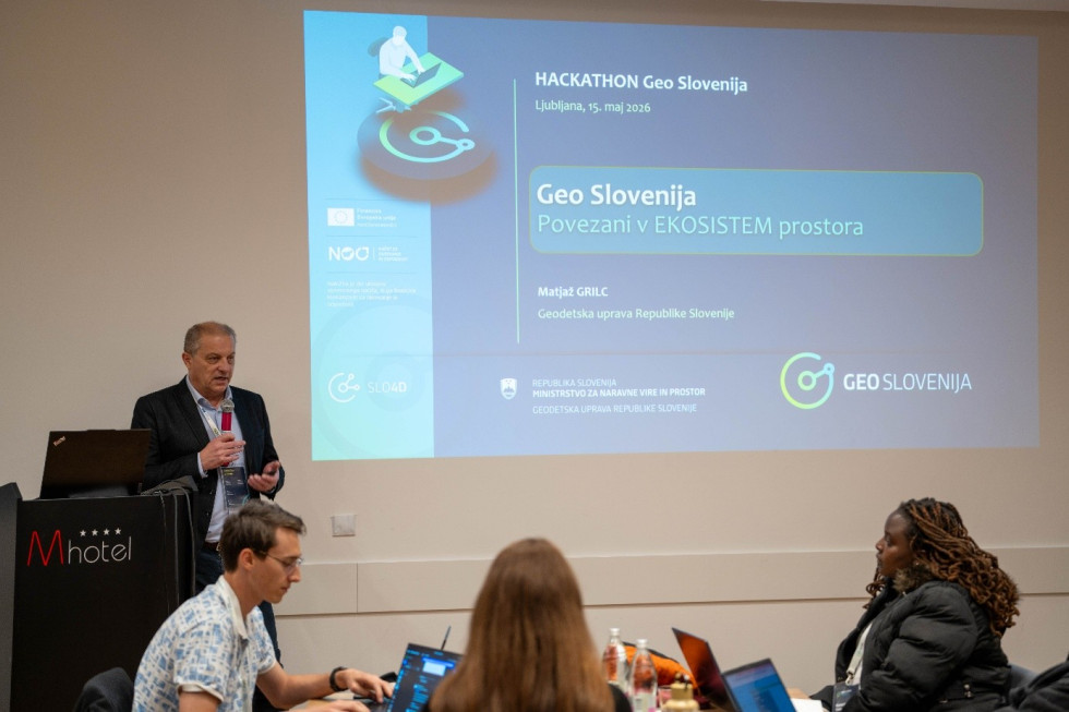 Moški v poslovni obleki stoji ob govorniškem pultu in govori v mikrofon. Za njim je projekcija z naslovom Hackathon Geo Slovenija, pred njim pa sedijo udeleženci z odprtimi prenosnimi računalniki.