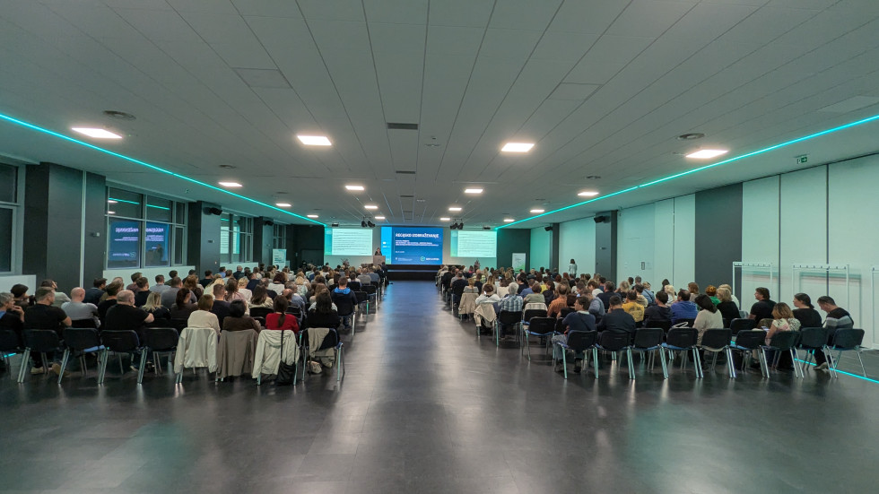 Velika skupina moških in žensk sedi v dvorani ter spremlja predstavitev na odru.