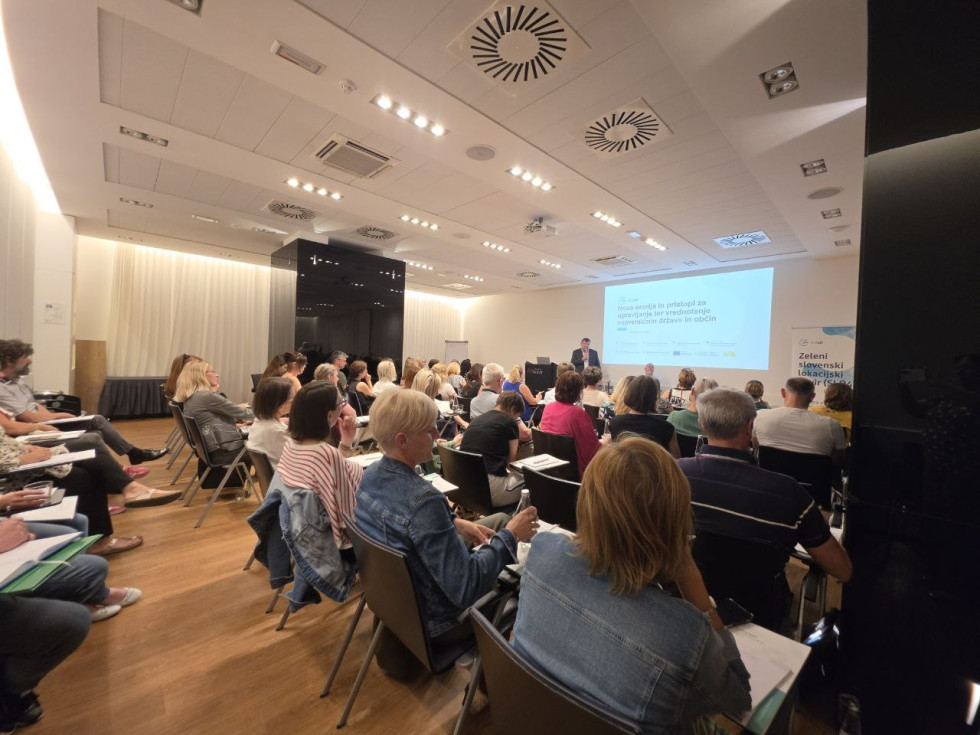Moški stoji pred udeleženci v konferenčni dvorani in predstavlja nova orodja ter pristope za upravljanje in vrednotenje nepremičnin države in občin.