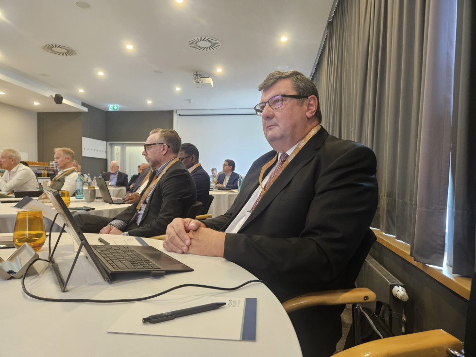 Moški v poslovni obleki sedi za okroglo mizo v konferenčni dvorani in spremlja razpravo. Pred njim sta prenosni računalnik in gradivo dogodka, v prostoru pa sedijo tudi drugi udeleženci srečanja.