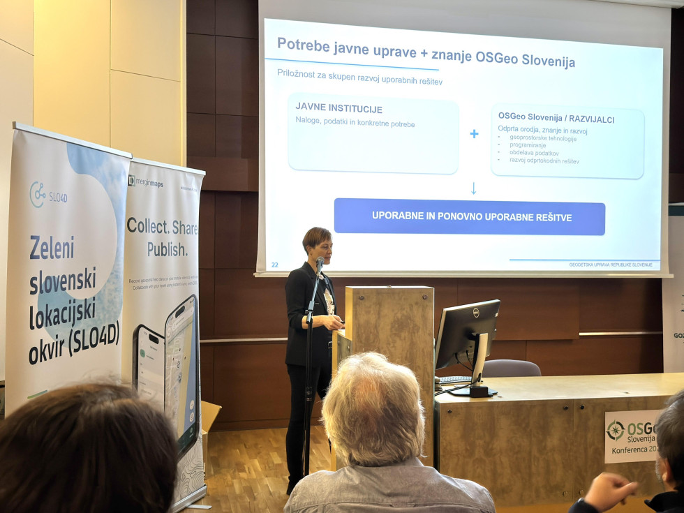Poslovno oblečena ženska za govorniškim pultom predstavlja povezovanje potreb javne uprave in znanja skupnosti OSGeo Slovenija za razvoj uporabnih rešitev.