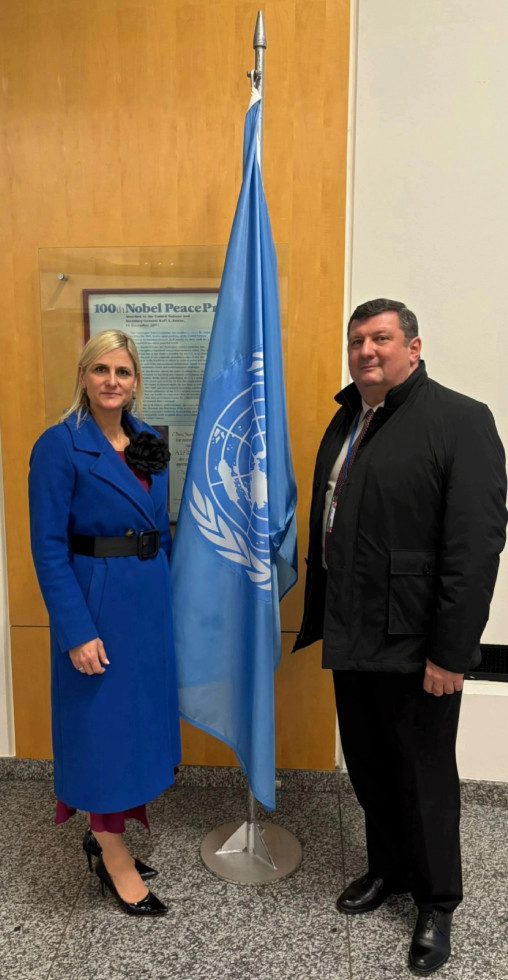 Ženska in moški stojita ob zastavi Združenih narodov v prostorih OZN na Dunaju; predstavljata Slovenijo na delavnici o integraciji geoprostorskih in statističnih podatkov.