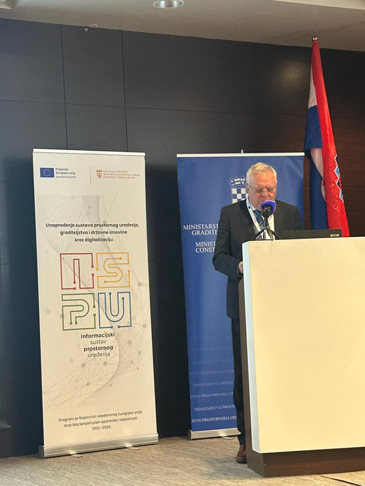 Na sliki stoji moški v poslovnih oblačilih. Moški stoji za govorniškim pultom, zadaj sta vidna dva panoja, na katerih so logotipi konference