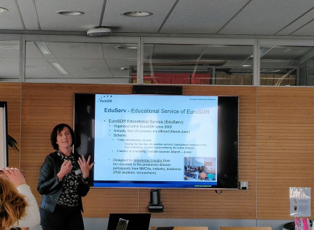 Ženska predavateljica stoji pred zaslonom z diapozitivom o izobraževalni službi EuroSDR, v sejni sobi za mizo sedijo udeleženci z odprtimi prenosniki.
