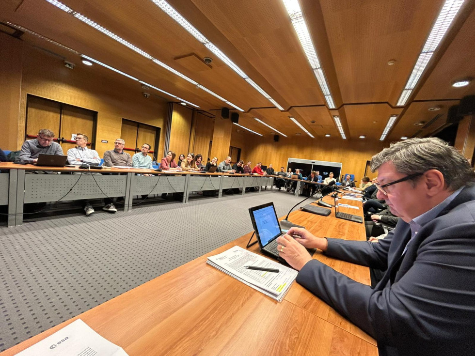 Moški sedi za mizo in uporablja tablico, v ozadju pa večja skupina udeležencev sedi v konferenčni dvorani in spremlja razpravo.