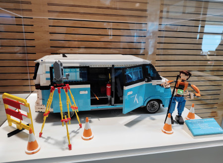 Maketa geodetskega kombija in figurice geodeta iz lego kock z merilnim stojalom ter prometnimi stožci v razstavni vitrini.