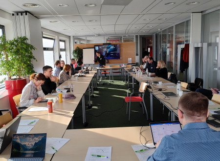 Skupina moških in žensk sedi za dolgimi mizami v sejni sobi, na koncu prostora poteka predstavitev na zaslonu, udeleženci uporabljajo prenosnike in zapiske.