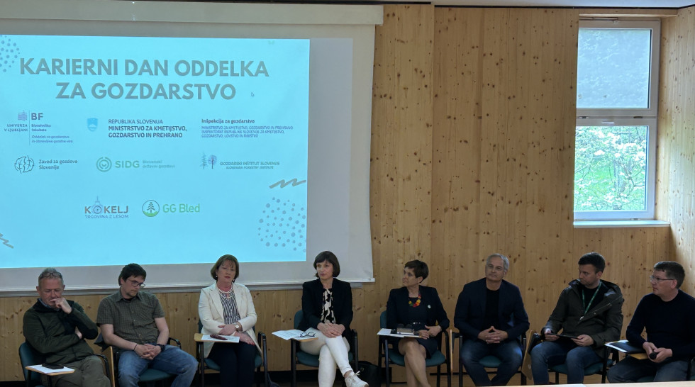 Ženske in moški sedijo na stolih. V ozadju na platnu predstavitev.