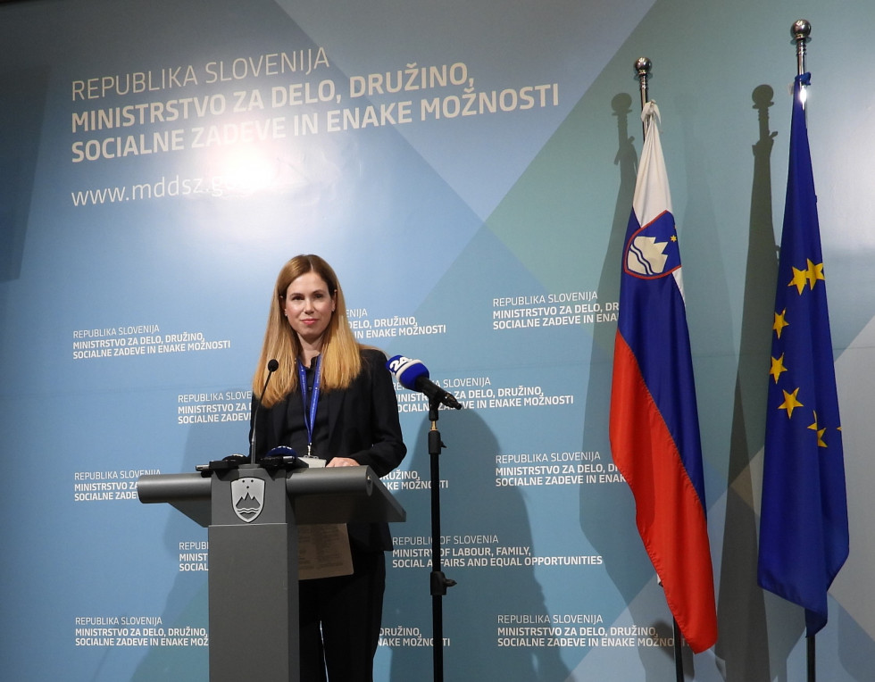 Ženska stoji za govorniškim pultom, za njo moder pano z napisi Republika Slovenija Ministrstvo za delo, družino, socialne zadeve in enake možnosti, ob strani slovenska in evropska zastava