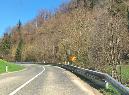 cesta v gozdu, ki ima na levi strani varovalni podporni zid, ki ima nameščeno varnostno ograjo za motoriste. Slika prikazuje levi ovinek in začasni prometni znak 70 km/h