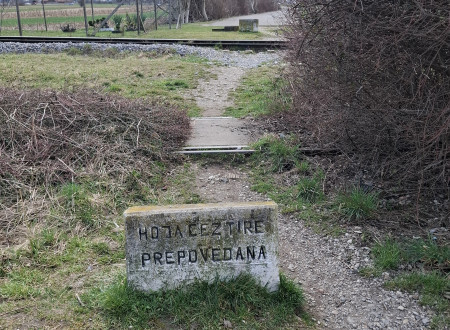 Nezavarovan prehod železniških tirov. Ob peščeni potki je postavljen kamen z vklesanim napisom "Ne hodi čez progo", ki pa v nadaljevanju potka prečka železniške tire.