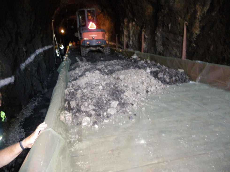 V temi tunela je osvetljen odprt železniški vagon, prekrit s plastično folijo, na katero stroj, viden v ozadju nalaga odpadni izkopni material s tal