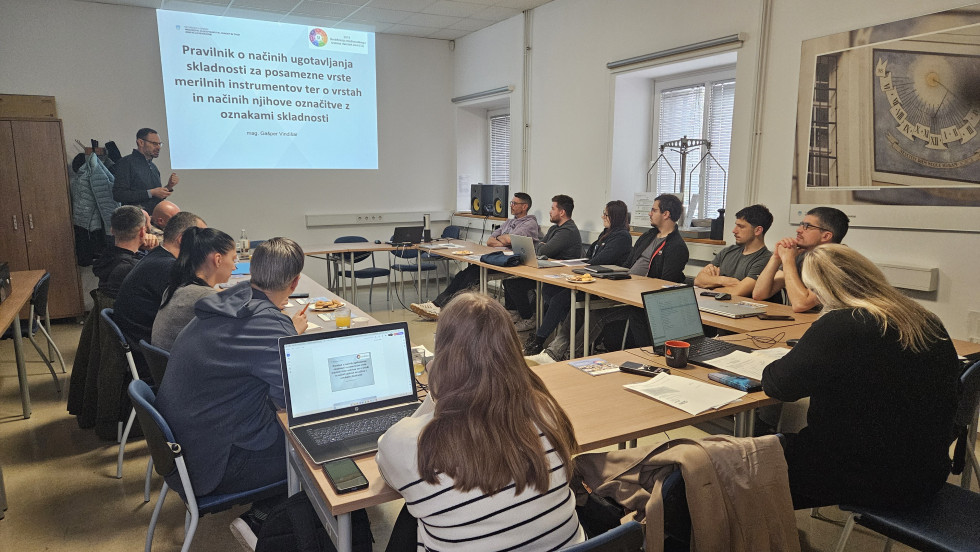 Na sliki je sejna soba z udeleženci, ki sedijo v kvadratu in poslušajo predavatelja spredaj.