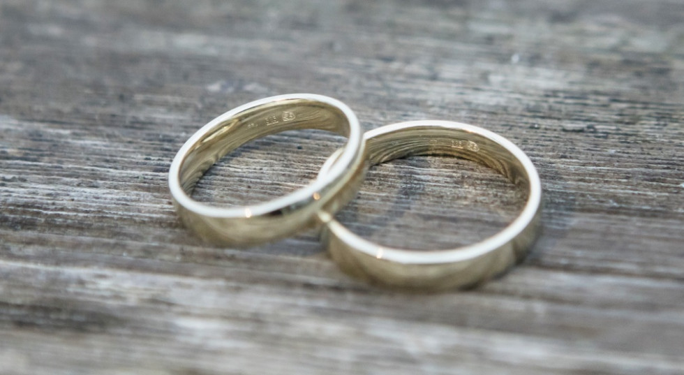 Two gold wedding rings are crossed on a wooden surface. Three assay marks are blurred on the rings.