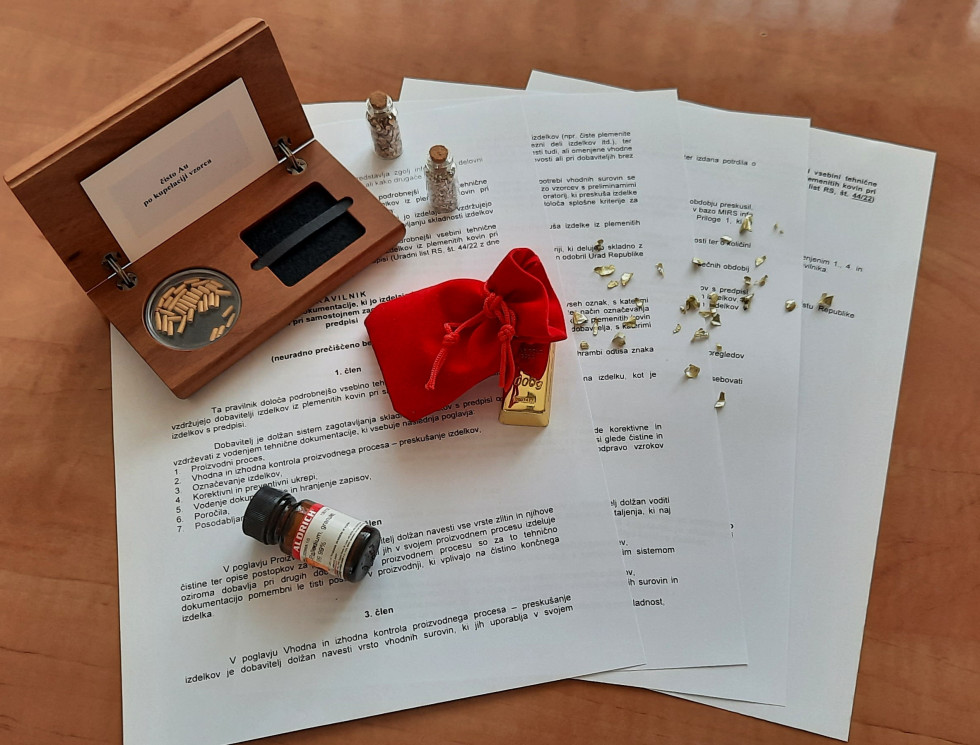 Na mizi so listi pravilnika, na njih pa lesena škatlica, rdeč mošniček in stekleničke s plemenitimi kovinami (zlato, srebro in paladij).