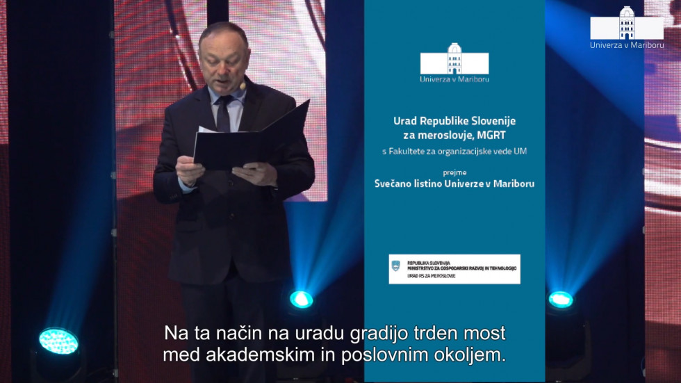 Na levi strani slike je povezovalec svečane prireditve, na desni strani pa modra pasica na kateri je zgoraj logo tip Univerze v Mariboru in spodaj Urada RS za meroslovje.