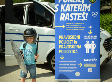 Otrok v opremi pravosodnih policistov, v ozadju službeno vozilo in plakat