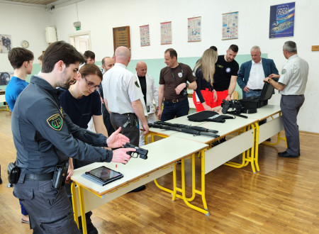 Pravosodni policist prikazuje delovanje pištole, v ozadju obiskovalci v pogovoru s pravosodnimi policisti