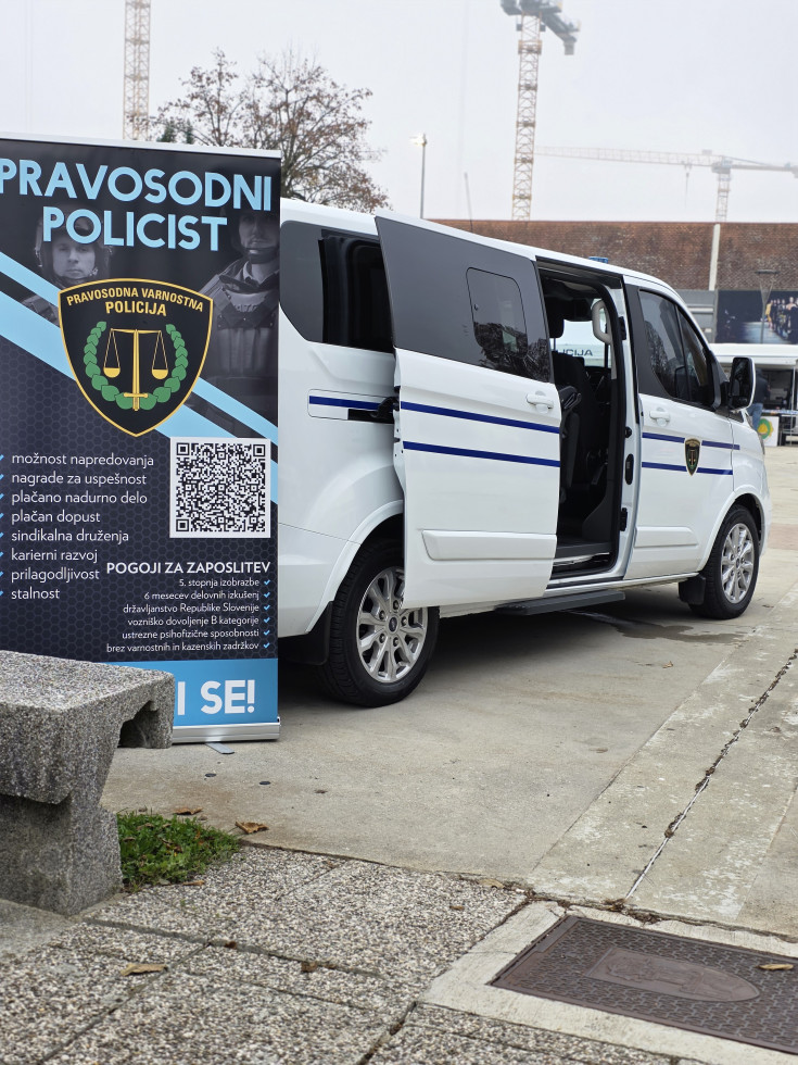 Na sliki je kombi, ki je oblepljen z dvema modrima črtama in znakom Uprave za izvrševanje kazenskih sankcij. V notranjosti je opremljen za prevoz zaprtih oseb.