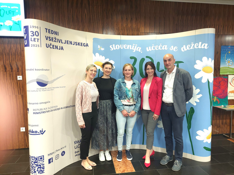 Skupinska fotografija zaposlenih iz ženskega zapora z obsojenko na sredini, v ozadju modro-bel pano vseživljenjskega učenja